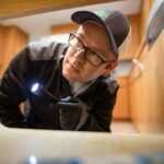 A person wearing glasses, a cap, and gloves is inspecting under a cabinet with a flashlight in a kitchen.