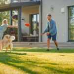 A family of four, along with a golden retriever, is playing frisbee on a lawn in front of a modern house. The sun is shining, and trees are visible in the background, providing natural mosquito control. The family members appear to be enjoying the outdoor activity together.