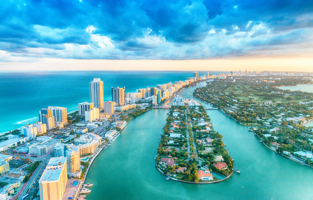 Aerial view of Miami's coastal cityscape where tall buildings stretch along the Florida shoreline, with a wide river running parallel. Residential areas dot both sides of the water, while a partly cloudy sky allows sunlight to illuminate the vibrant scene.