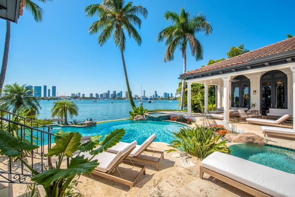 A luxurious poolside scene in Miami featuring a waterfront view with palm trees, a swimming pool, and lounge chairs. A spacious patio is bordered by tropical plants, and Florida's vibrant city skyline is visible across the water under a clear blue sky.