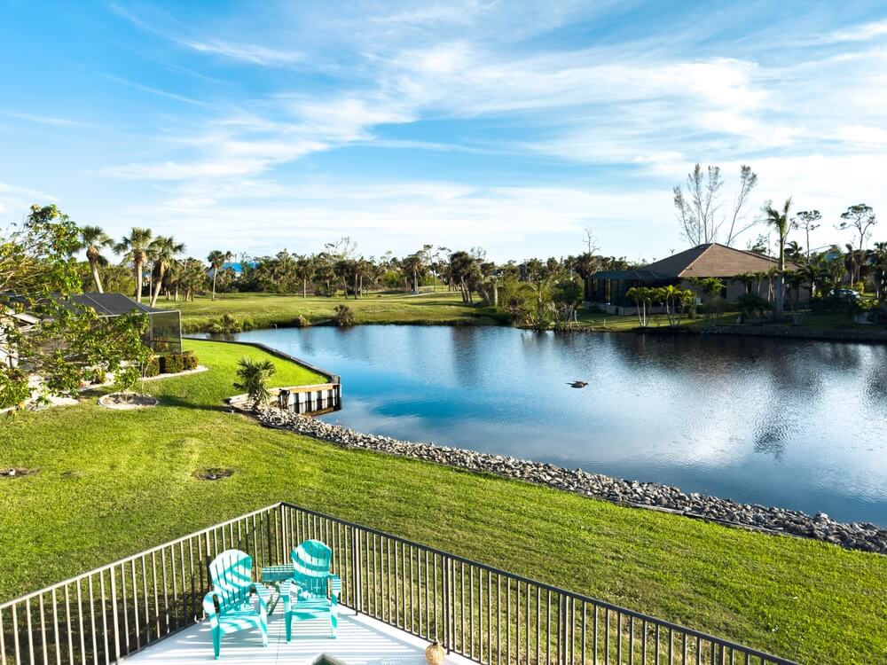 A tranquil Miami scene features a small lake with a house on either side, surrounded by lush green lawns. Two turquoise chairs sit on a patio overlooking the water. The sky is clear with scattered clouds, and trees line the background in this peaceful corner of Florida.