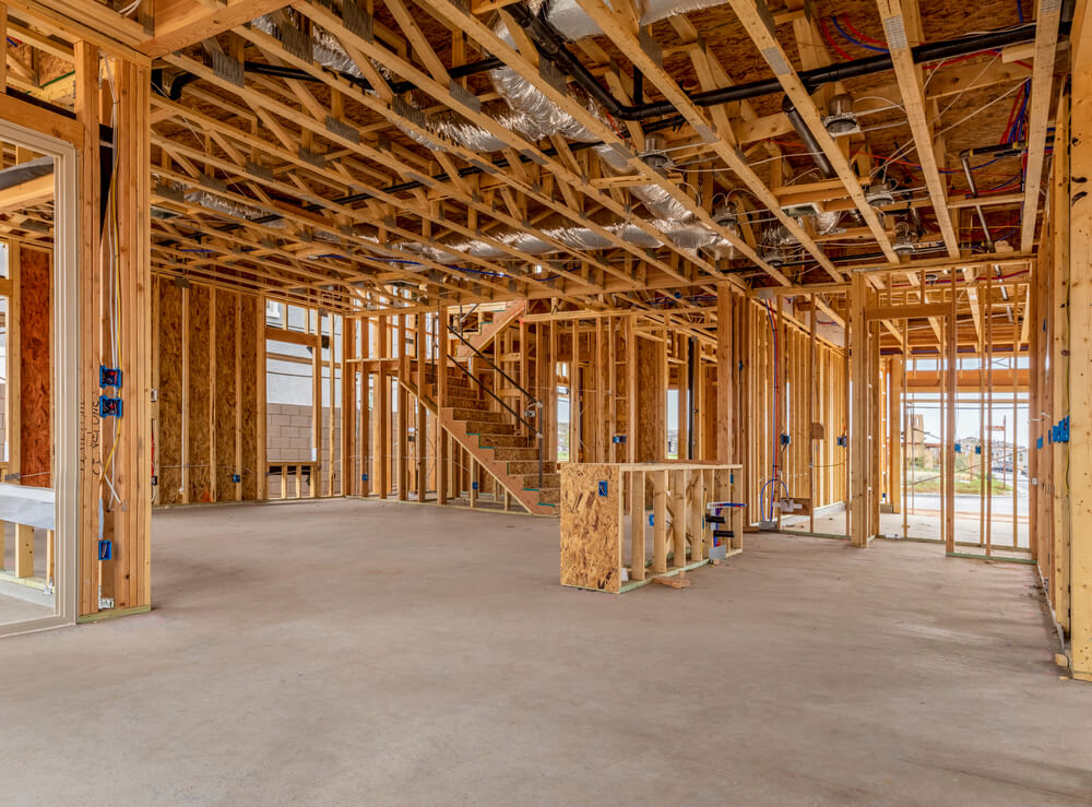 A construction site in Miami reveals the interior framework of a building. Wooden beams and studs outline the structure, with visible wiring and ductwork. An unfinished staircase leads to an upper level, and Florida's bright daylight streams through unglazed openings.