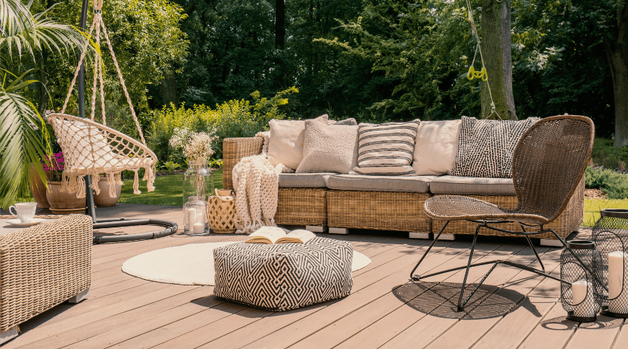 Outdoor patio with a wicker couch and cushions, a patterned ottoman, and a wire-frame chair on a wooden deck. A hanging chair is in the corner. Plants and lanterns decorate the area, creating an inviting escape for reading or enjoying summer pest control tips from the book open on the ottoman.