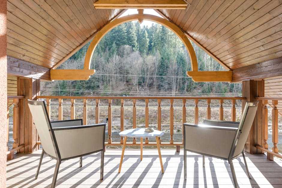 A wooden balcony with two modern chairs and a round table offers a view of distant trees under a clear sky, framed by a large arched window. Watch out for the occasional summer spider weaving its web in this serene retreat.