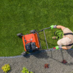 A person in gardening attire is pushing a red lawn mower over a well-manicured lawn, ideal for lawn aeration. The grass is bordered by a strip of gravel decorated with a few small plants. The scene is viewed from above.