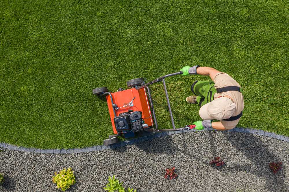 A person in gardening attire is pushing a red lawn mower over a well-manicured lawn, ideal for lawn aeration. The grass is bordered by a strip of gravel decorated with a few small plants. The scene is viewed from above.