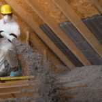 A worker in protective gear is expertly spraying pest control insulation inside an attic. The person wears a yellow hard hat, white coveralls, gloves, and a mask. The attic's wooden beams are visible, with insulation panels neatly positioned along the wall.