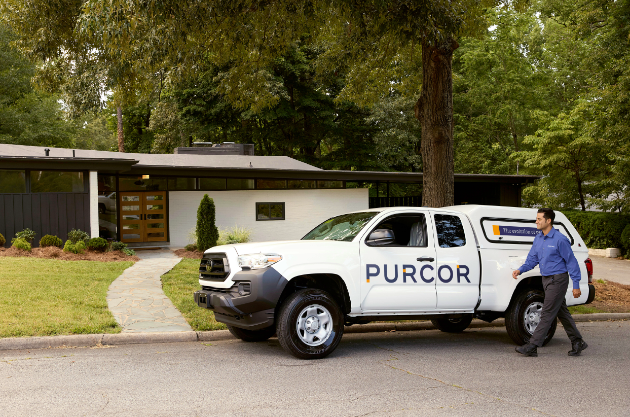 A man in a blue shirt walks towards a white pickup truck with "PURCOR" written on the side, offering top tips for wasp control. The truck is parked in front of a modern house surrounded by San Diego's lush trees and greenery.