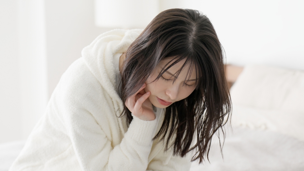 A woman with long dark hair wearing a white hoodie sits on a bed, appearing deep in thought or feeling unwell. Her head is tilted slightly downward, and her hand rests on her chin, possibly contemplating bug bite symptoms. The background is softly lit, adding to the contemplative mood.