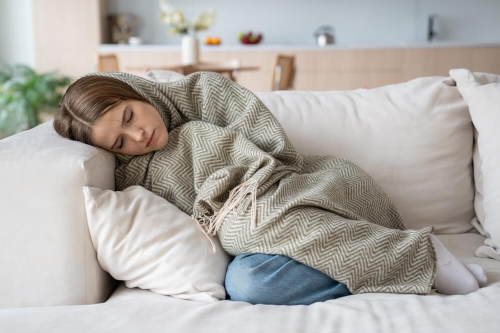 A person is peacefully sleeping on a beige couch, wrapped in a gray and white patterned blanket for a pest-free home. Their head rests on a pillow, and they appear comfortable. The background shows a blurred kitchen area with various items on the countertop, possibly in Brunswick GA.