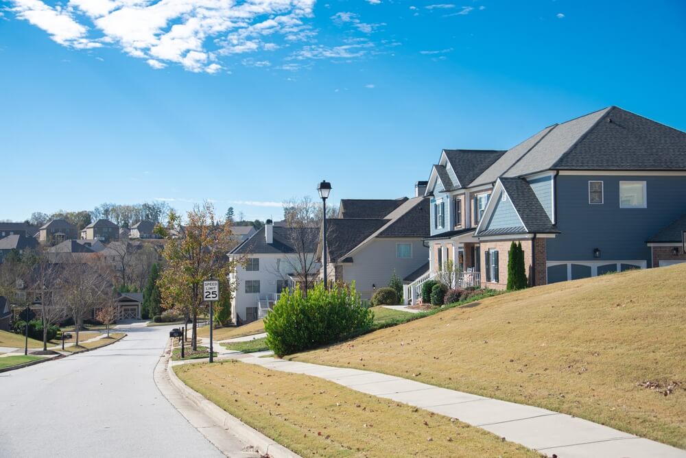 A suburban street in Brunswick, GA features neatly lined, pest-free homes under a clear blue sky. The road slopes gently with a visible 25 mph speed limit sign. Each house boasts well-maintained lawns, and the mostly bare trees hint at late autumn's embrace.