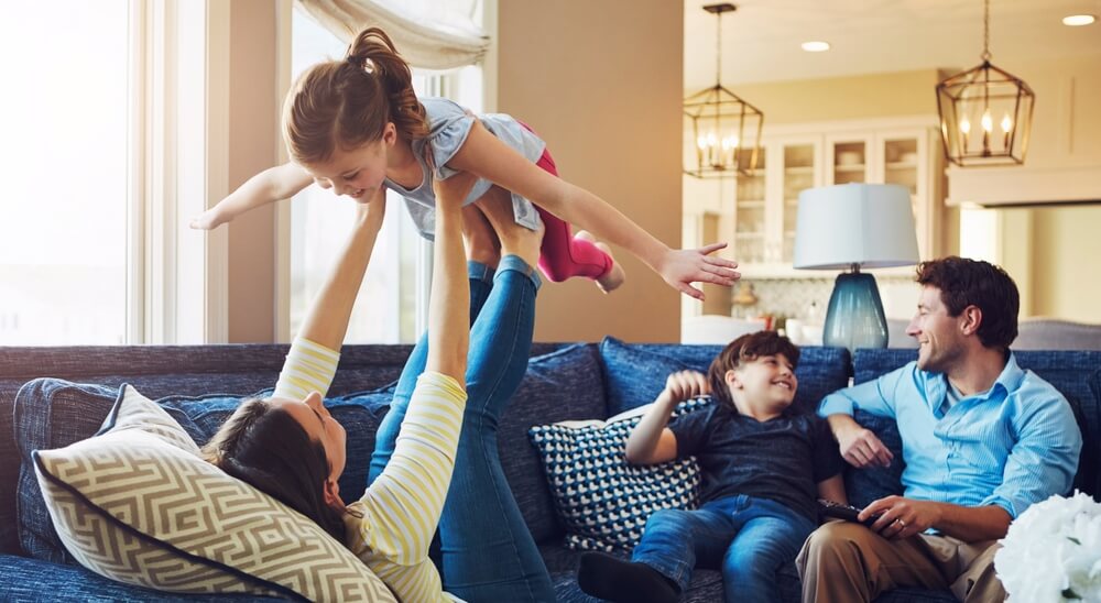 In their well-lit Brunswick, GA living room with modern decor and pendant lights, a family enjoys quality time together. A woman playfully lifts a young girl into the air on the sofa while a man and boy watch and smile, all cherishing their pest-free home environment.